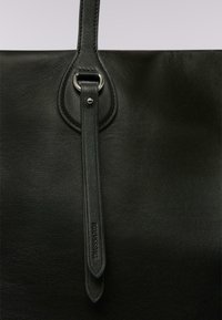Bolso de mano de cuero negro con una textura suave, un acento de herrajes circular y una larga correa plana de cuero con branding en relieve.