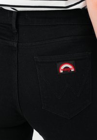 Gros plan sur la poche arrière d'un jean noir avec un patch logo Wrangler rouge et blanc, porté avec un haut rayé noir et blanc.