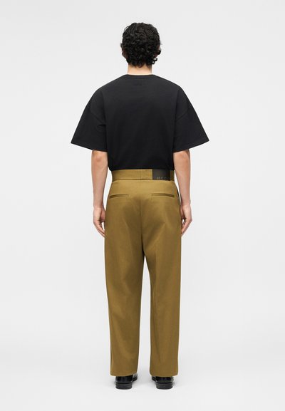 Homme aux cheveux bouclés portant une chemise noire à manches courtes rentrée dans un pantalon marron taille haute, debout de dos sur un fond blanc.