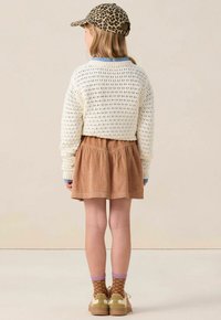 Enfant portant une casquette en imprimé léopard, un pull en tricot crème, une jupe en velours côtelé marron, des chaussettes à motifs et des baskets beiges, debout de dos sur un fond neutre.