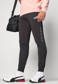 Zwarte sweatpants met wit Puma-logo, taps toelopende pasvorm, zijstrepen met logo-accenten, gecombineerd met zwart-witte sportschoenen.
