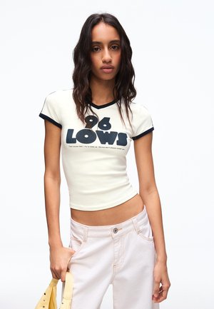 Mladá žena s dlhými tmavými vlasmi, oblečená v bielom krátkom topu s nápisom „96 LOWS“ a svetloružových nohaviciach, drží žltú kabelku po boku.