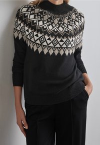Mörk tröja med hög hals och geometriskt mönster i ljusbrun och krämfärg över bröstet, med ribbade ärmslut och nederkant.