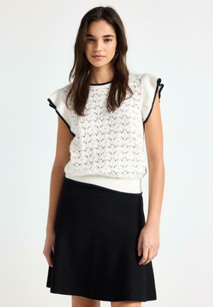 Ung kvinde med langt mørkt hår iført en hvid strikket ærmeløs top med sort kant og en sort knælængde nederdel foran en ensfarvet baggrund.