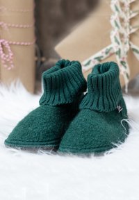 Chaussons en laine verte avec poignets côtelés posés sur un tissu en fourrure blanche, avec des cadeaux emballés et attachés avec des rubans en arrière-plan.
