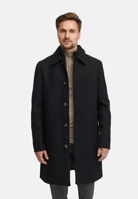 Svartullkåpe med krage, frontknapper og sidelommer. Båret over en beige høyhalset genser og grå bukser.