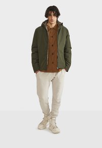 Giacca imbottita verde scuro con cappuccio, chiusura con zip e interno foderato, indossata sopra una camicia marrone a bottoni e pantaloni jogger beige.