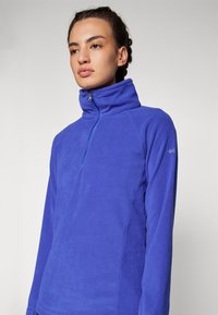 Ljubičasti flis pulover s visokim ovratnikom i pola-zip prednjim zatvaranjem. Ima duge rukave i suptilan detalj s logotipom na lijevoj ruci.