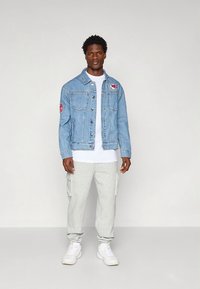 Gaiši zila džinsa jaka ar priekšējiem kabatām un logotipa uzliku, kopā ar baltu T-kreklu, pelēkām cargo biksēm un baltām sporta kurpēm.