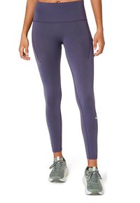 Femme portant des leggings de sport violets taille haute avec poches latérales et des chaussures de course grises, se tenant contre un fond blanc.