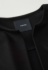 Vêtement noir au toucher doux avec un col rond. Comprend une étiquette en tissu avec le mot "un jour." en texte blanc.