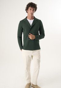 Giacca doppiopetto verde con texture e colletto a scialle, abbinata a pantaloni beige e scarpe chiare. Canottiera bianca semplice visibile.