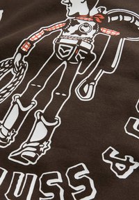 Tessuto marrone con un design grafico di un cowboy con un lazo, accenti in camicia a quadri rossi e contorni bianchi. Testo sotto l'immagine.