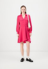 Abito avvolgente rosa con maniche lunghe, due tasche frontali e vita con cintura. Indossato con mocassini neri e calze crema, su uno sfondo bianco.
