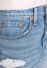 Světle modré džíny z denimu mají distressed design s třepícími se okraji, měděnými nýty, vybledlou texturou a klasickým pětiokýnkovým stylem.