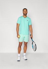 Mint green athletic t-shirt with short sleeves and contrasting seam accents, paired with matching shorts. Holding a tennis ball and racquet.
