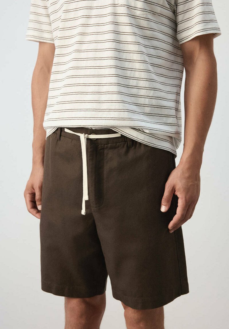 Homme portant un short marron foncé avec un cordon blanc et une chemise blanche à manches courtes avec de fines rayures horizontales marron.