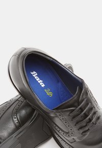 Zapatos de vestir de cuero negro con diseño de brogue, acabado suave y plantilla azul con el logo "Bata 24h". Forma elegante y formal.