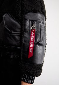 Schwarze Leder- und Kunstfell-Bomberjacke mit einer Reißverschlusstasche und einem roten "REMOVE BEFORE FLIGHT"-Tag, der am Ärmel angebracht ist. Strukturiertes Material.