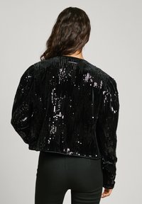 Musta samettinen lyhyt takki, jossa on paljettikoristelu, puffatut hartiat ja sileä pinta takana, yhdistettynä slim fit mustiin housuihin.
