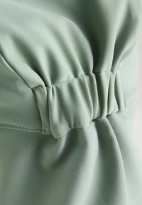 Podrobnost svetlo zelene obleke s shranjenim pasom z elastiko in gladko teksturo, ki prikazuje urejene šive in mehko padajoč material.
