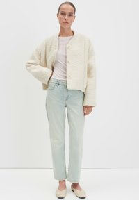 Cremfarbige, strukturierte Jacke mit cropped Design und rundem Ausschnitt, kombiniert mit hellblauen, geraden Jeans und beigen Slippern.