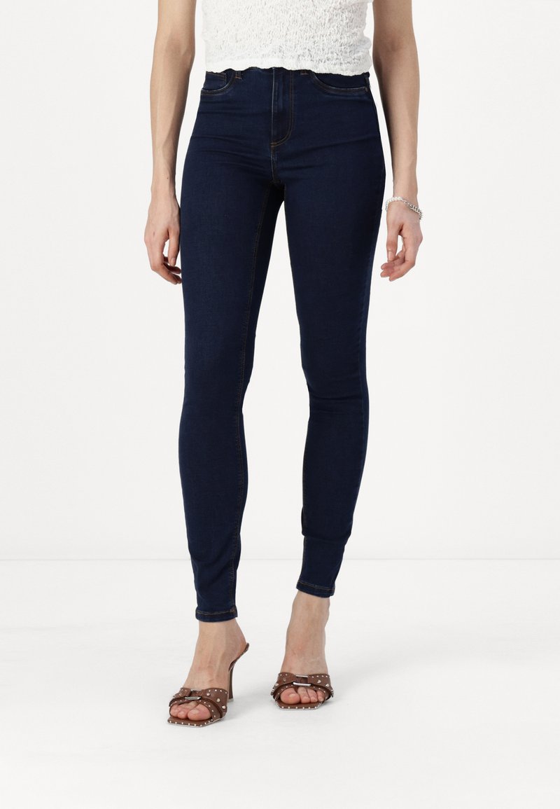 Skinny dark blue high-waisted jeans with a smooth texture and contrasting stitching. Paired with brown studded slide sandals.
