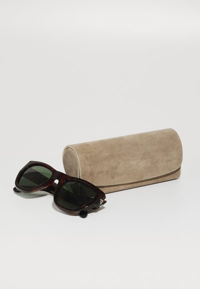 Óculos de sol com lentes verde-escuro e armações de tartaruga em padrão castanho, ao lado de uma capa de camurça bege com um logo discreto.