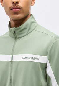 Άνδρας που φοράει ανοιχτοπράσινο μπουφάν με φερμουάρ, με λευκή οριζόντια ρίγα και το κέντημα «J.LINDEBERG» στο στήθος.