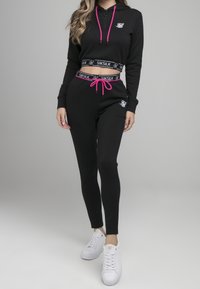 Ensemble de sport noir comprenant un sweat à capuche court et un pantalon ajusté. Comprend des cordons de serrage roses et des accents de logo. Des baskets blanches complètent le look.