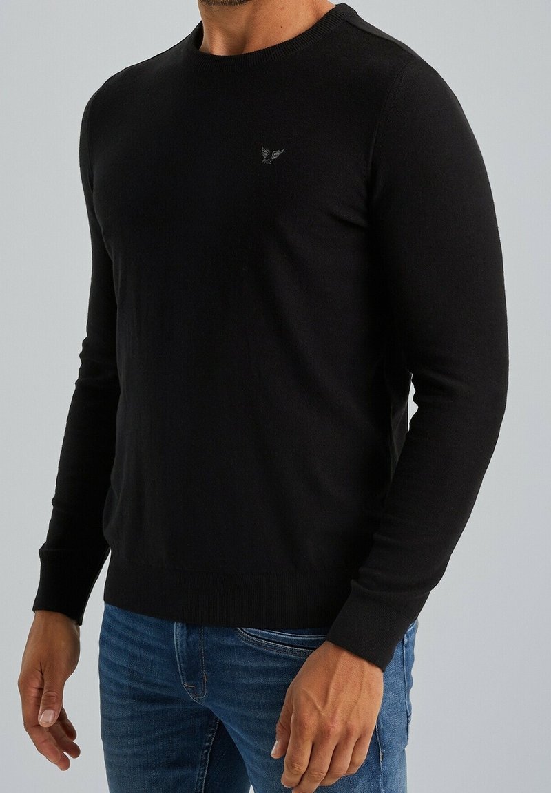 Homme portant un pull noir ajusté à manches longues avec un petit emblème sur la poitrine et un jean bleu sur un fond uni.