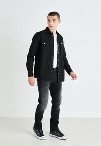 Black denim shirt with snap buttons, white t-shirt underneath, paired with dark grey jeans and black high-top sneakers. Neutral background.