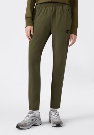 Osoba ubrana w oliwkowozielone spodnie sportowe z czarnym logo oraz szare tenisówki z białymi sznurowadłami i skarpetkami, stojąca na białym tle.