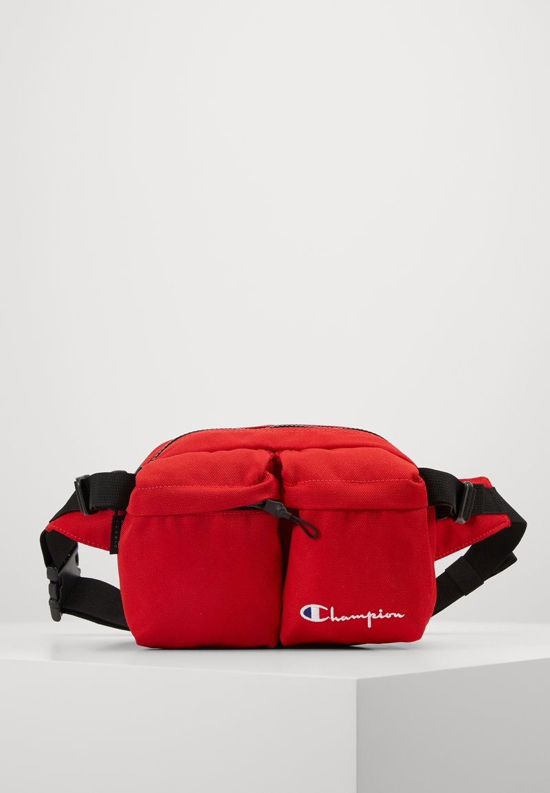 Sac banane rouge Champion avec deux poches zippées à l'avant et une sangle noire réglable sur une surface blanche unie.