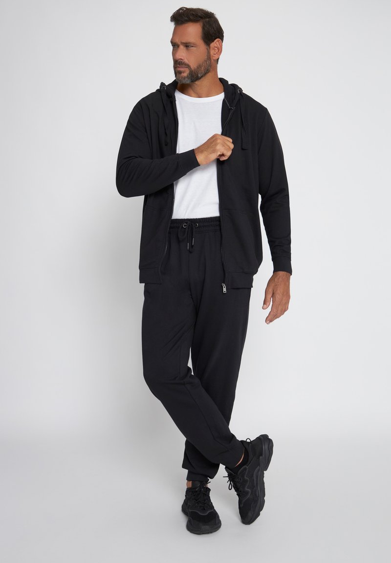 Man wearing black zip-up hoodie, black jogger pants, black sneakers, and white t-shirt, standing with one leg crossed in studio.