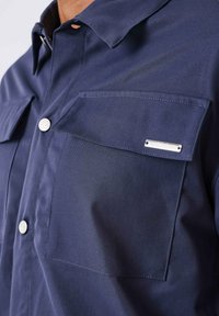 Navyblauw shirt van gladde stof, met een knoopsluiting aan de voorkant, twee borstzakken en een metalen merklabel op de rechterzak.
