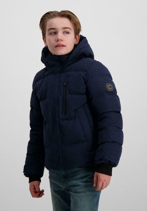 Een jonge persoon in een marineblauwe gewatteerde jas met capuchon en jeans kijkt over zijn schouder tegen een effen achtergrond.