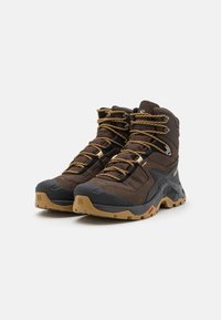 Brązowe buty outdoorowe z teksturowaną górną częścią tkaniny, gumowym czubkiem i mocną podeszwą. Posiadają żółte sznurówki i metalowe oczka dla pewnego zapięcia.