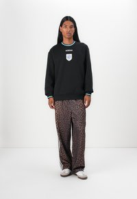 Sort sweatshirt med et Adidas-logo og mærkedetalje, parret med bukser i brunt leopardprint. Hvide sneakers med grå accenter.