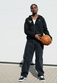 Sudadera con cremallera negra con logo, camiseta blanca, jeans holgados negros y zapatillas negras, sosteniendo un balón de baloncesto, contra un fondo simple.