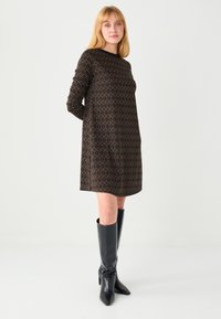 Robe marron à motifs géométriques, manches longues et coupe droite. Portée avec des bottes en cuir noir montant jusqu'aux genoux.