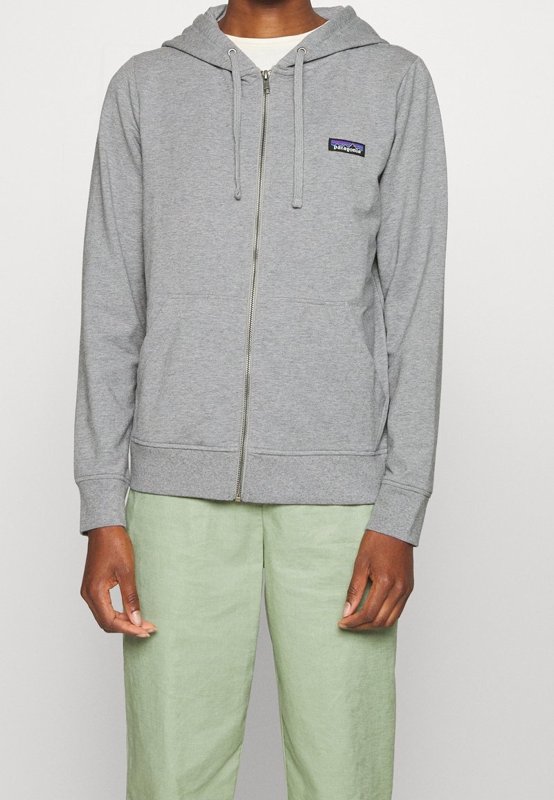 Personne portant un sweat à capuche zippé gris clair avec un patch logo Patagonia et un pantalon vert clair, debout devant un fond uni.