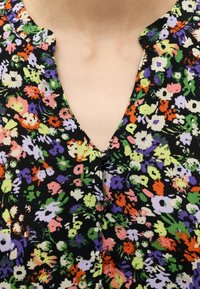 Prim-plan al unei bluze negre cu un model floral colorat în violet, portocaliu, verde, alb și roz, având decolteu în V și nasturi.