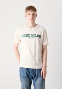 Cream cotton t-shirt with a crew neck, featuring green graphic text "GANT NH. CT. SPORTS CLUB" on the front, paired with blue denim jeans.