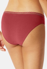 Rød bomuldsbikini med en bølget blondekant om taljen. Stoffet har en glat tekstur, der fremhæver et tætsiddende design uden ekstra pynt.