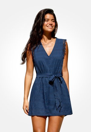 Donna sorridente con lunghi capelli scuri che indossa un vestito blu navy senza maniche con rifiniture in pizzo e cintura legata in vita, in piedi su sfondo bianco.