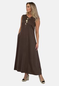 Brązowa, bezrękawnikowa sukienka maxi z gładkiego materiału, z centralnym szwem i złotą naszyjnikiem w formie wisiorka jako detal. Modelka nosi espadryle.