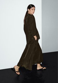 Long olive green satin dress with wide sleeves, featuring a relaxed fit and side pockets, paired with black flip-flops.