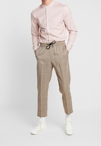 Ljusrosa långärmad skjorta med ståkrage och ficka, i kombination med brunrutiga tapered-byxor och vita träningsskor.