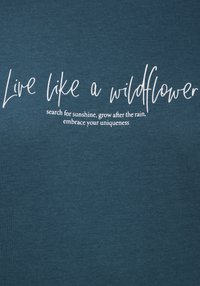 Teal bomull t-shirt med vit text som säger "Lev som en vildblomma" och ett litet inspirerande meddelande nedanför.